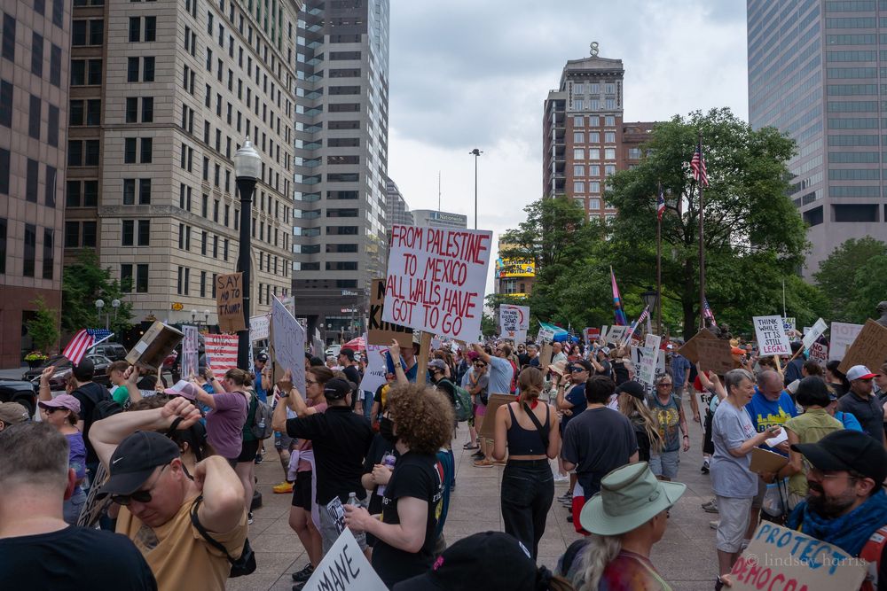 Crowd of protesters gather in downtown city area holding signs and marching past tall office buildings.