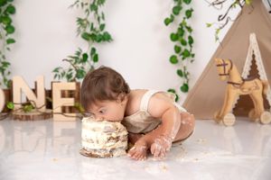 Baby wearing white dress dives face first into birthday cake next to decorative teepee and vine decor.