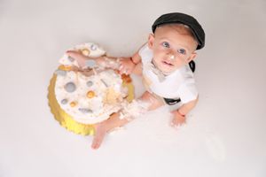 Baby in white outfit and cap smiling while playing with smashed cake on white background.