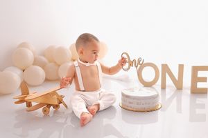 Baby in white outfit sitting next to wooden airplane toy and ONE sign with white balloon backdrop.