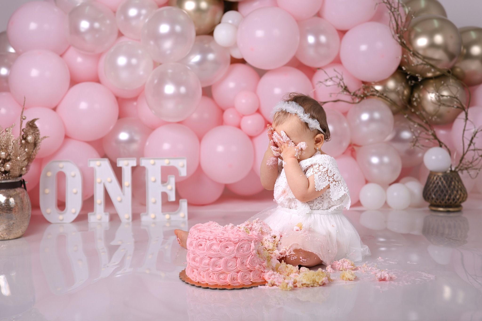 Baby in white tutu dress with pink and pearl balloon backdrop celebrating first birthday.
