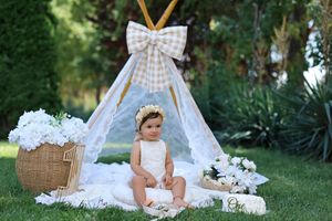 Baby in white romper sits on blanket before lace teepee with bow, surrounded by white flowers and wicker basket outdoors.