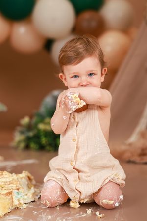 Baby in beige romper enjoying cake smash photoshoot with balloons.