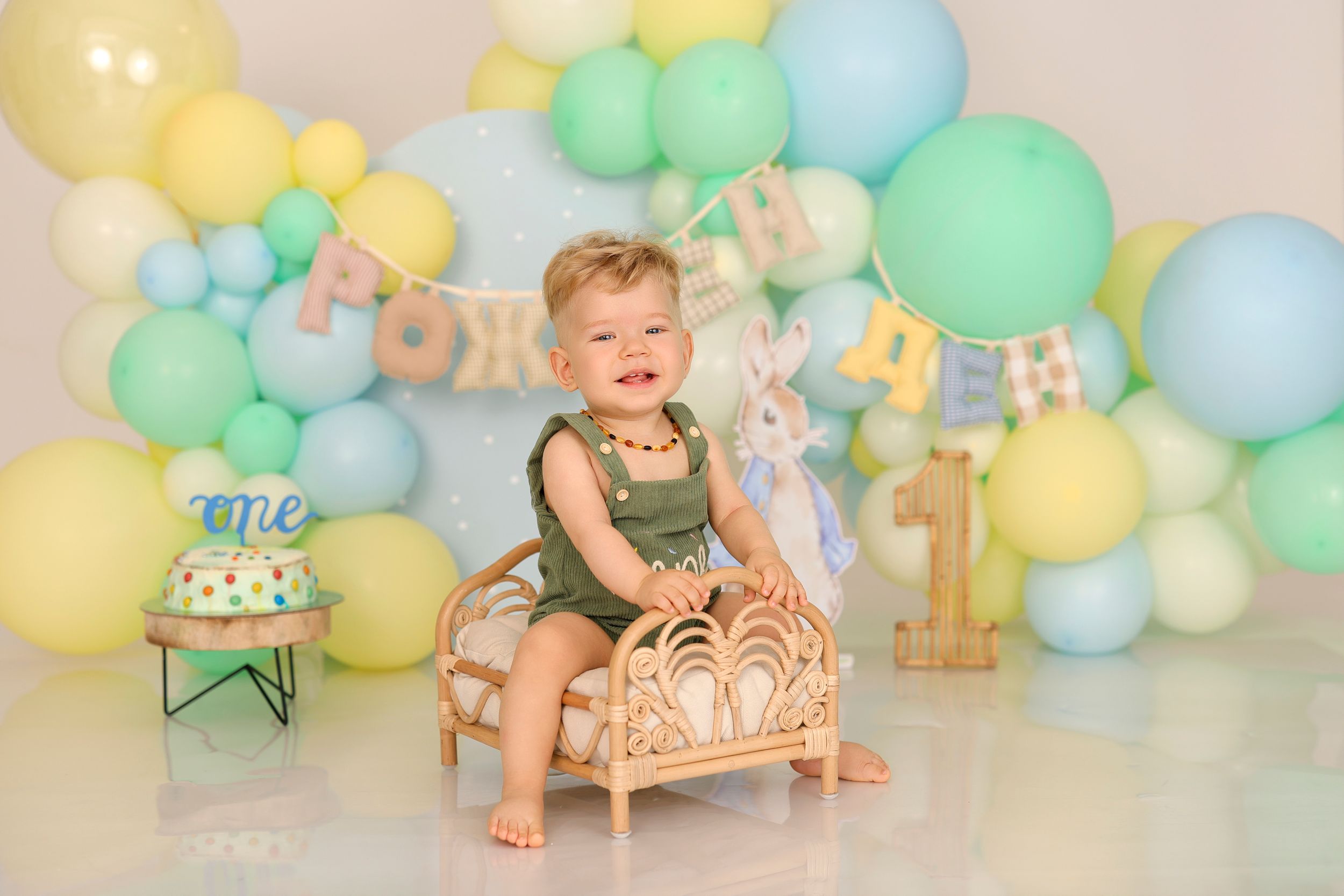 Smiling baby boy in green romper sits on rattan chair surrounded by pastel balloons and Easter decorations for first birthday.