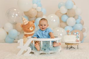 Baby boy in blue outfit sits at first birthday cake smash setup with balloons, teddy bear, and number 1 decor.