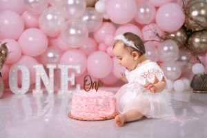 Baby girl in tutu dress celebrates first birthday with pink cake and balloon display.