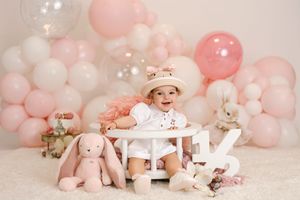 Baby girl smiles at her first birthday cake smash surrounded by pink balloons and stuffed bunnies.