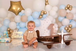 Baby boy smiles at first birthday party with balloon garland, wooden airplane, and Happy Birthday neon sign.