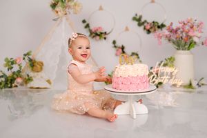 Baby girl in pink dress smiles at first birthday cake smash with floral boho backdrop.