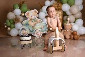 Baby boy in bowtie poses with toy scooter at teddy bear first birthday cake smash with green and gold balloon backdrop.