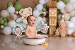 Happy baby boy sits in wash basin surrounded by teddy bear balloons and ONE blocks for first birthday photos.