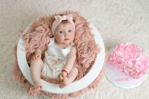 Baby girl in white dress with pink bow headband lies in bowl on fluffy pink fur next to first birthday cake.