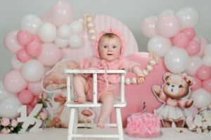 Baby girl in pink outfit sits in white high chair surrounded by pink balloons and teddy bear decorations at first birthday.
