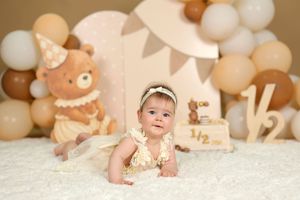 Baby girl in a cream dress crawls on a white rug at a half birthday party with bear and balloon decorations.