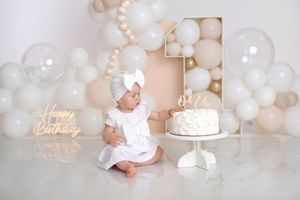 Baby girl in white dress sits beside a smash cake at her first birthday party with balloon decorations.