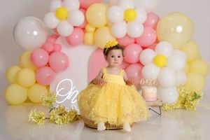 Baby girl in yellow lace dress sits in wooden bowl surrounded by pink, white, and yellow balloon arch backdrop.