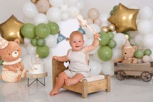 Baby sitting on a wooden bed surrounded by green, white, and gold balloons with a bear cake and toy train for a first birthday.