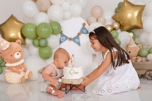 A baby enjoys their first birthday cake smash with an older sister, surrounded by green and gold balloon decor.