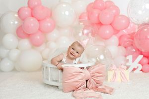 Baby girl in pink birthday hat sits in white crib surrounded by pink and white balloons with gifts.