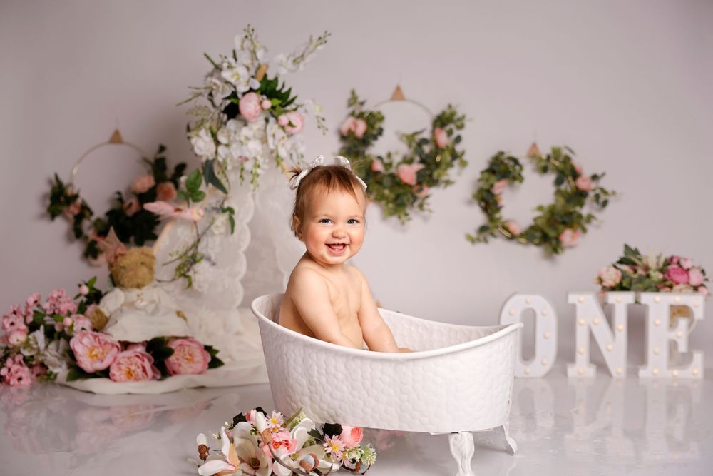 Floral themed cake smash photo setup with pink roses, white decorations and bathtub prop against neutral backdrop.