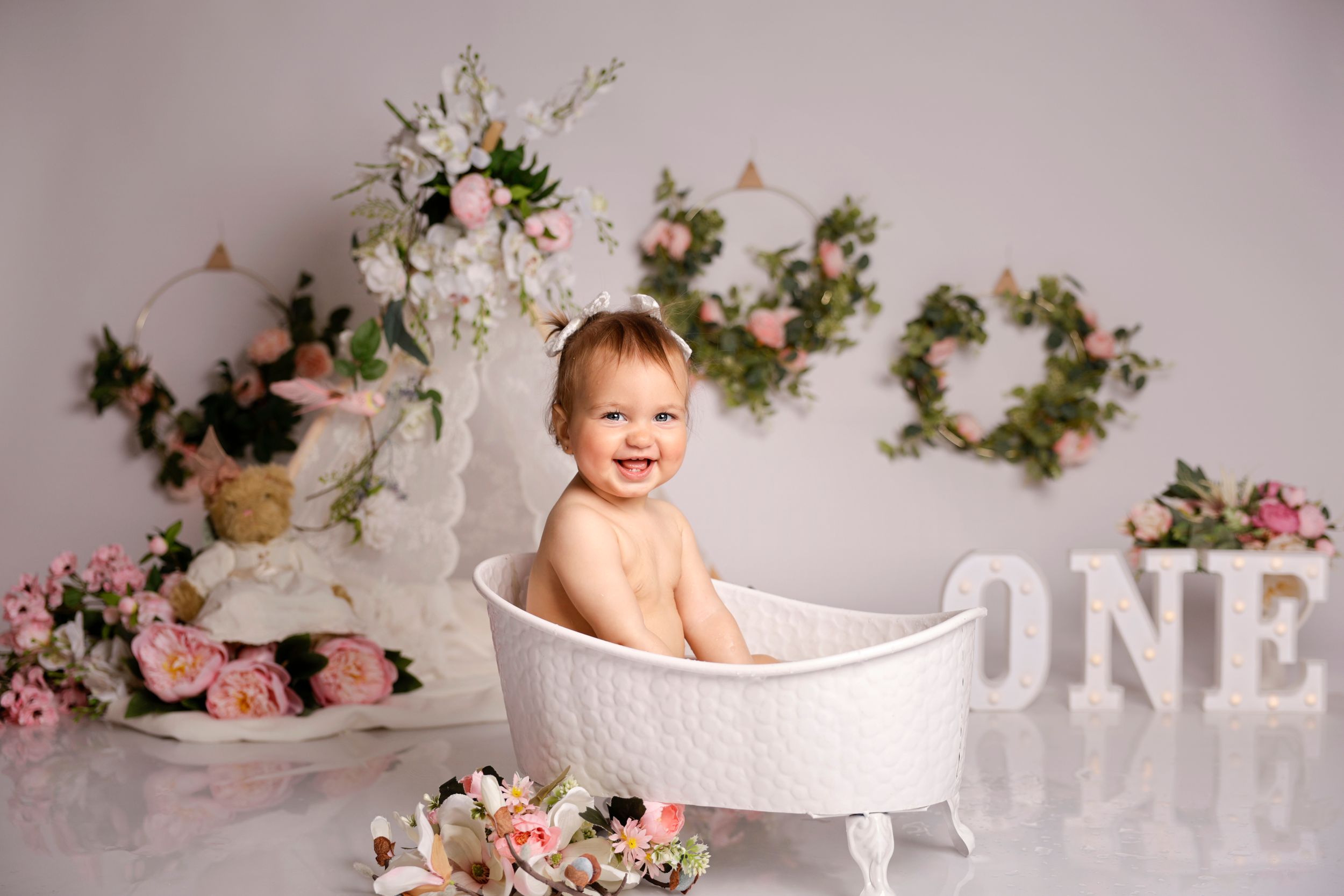 Floral themed cake smash photo setup with pink roses, white decorations and bathtub prop against neutral backdrop.