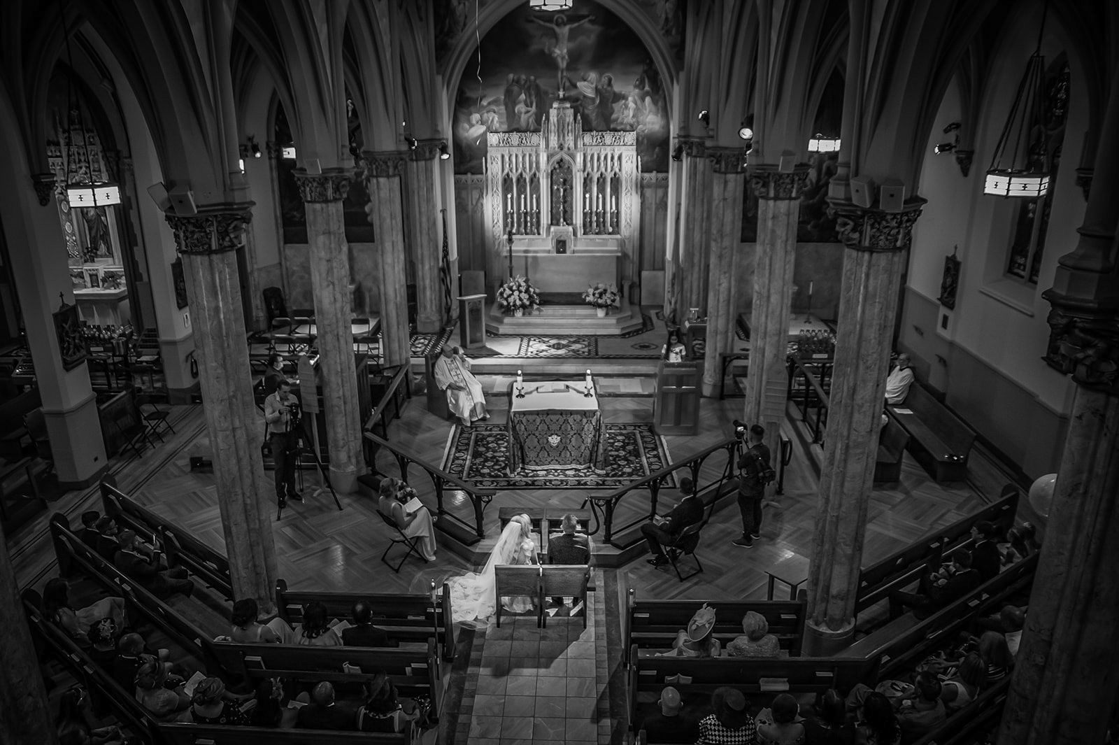 High-angle black and white view of a wedding ceremony inside an ornate cathedral with guests seated in curved pews.