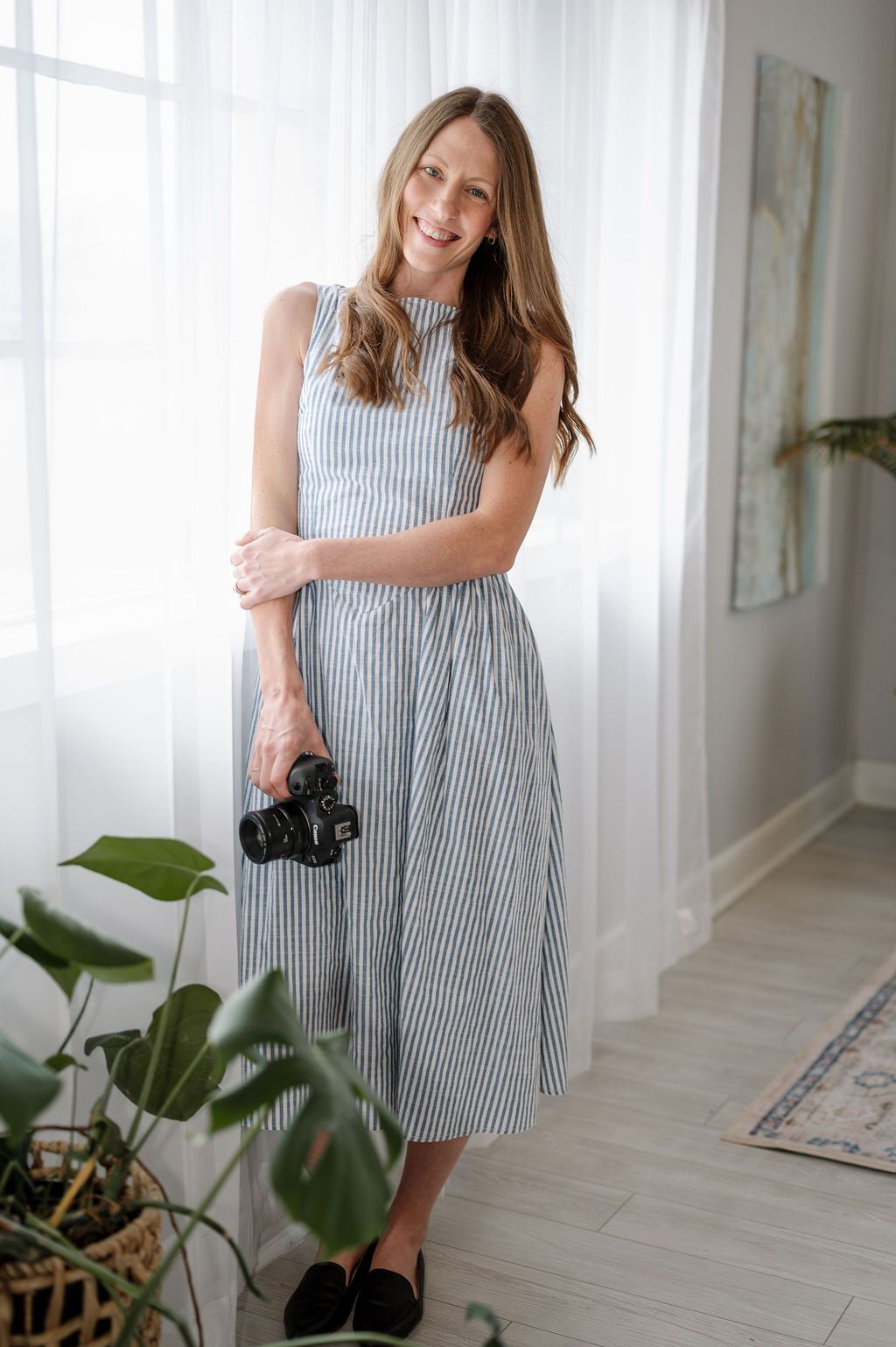 A photographer in a striped dress holds a camera in a bright, modern interior. Studio headshots