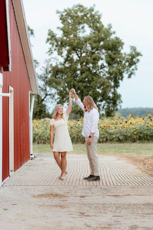 Couple in white clothing walking together near a red barn with sunflowers in the background. Mount Pleasant Michigan Heirloom Grove Venue.