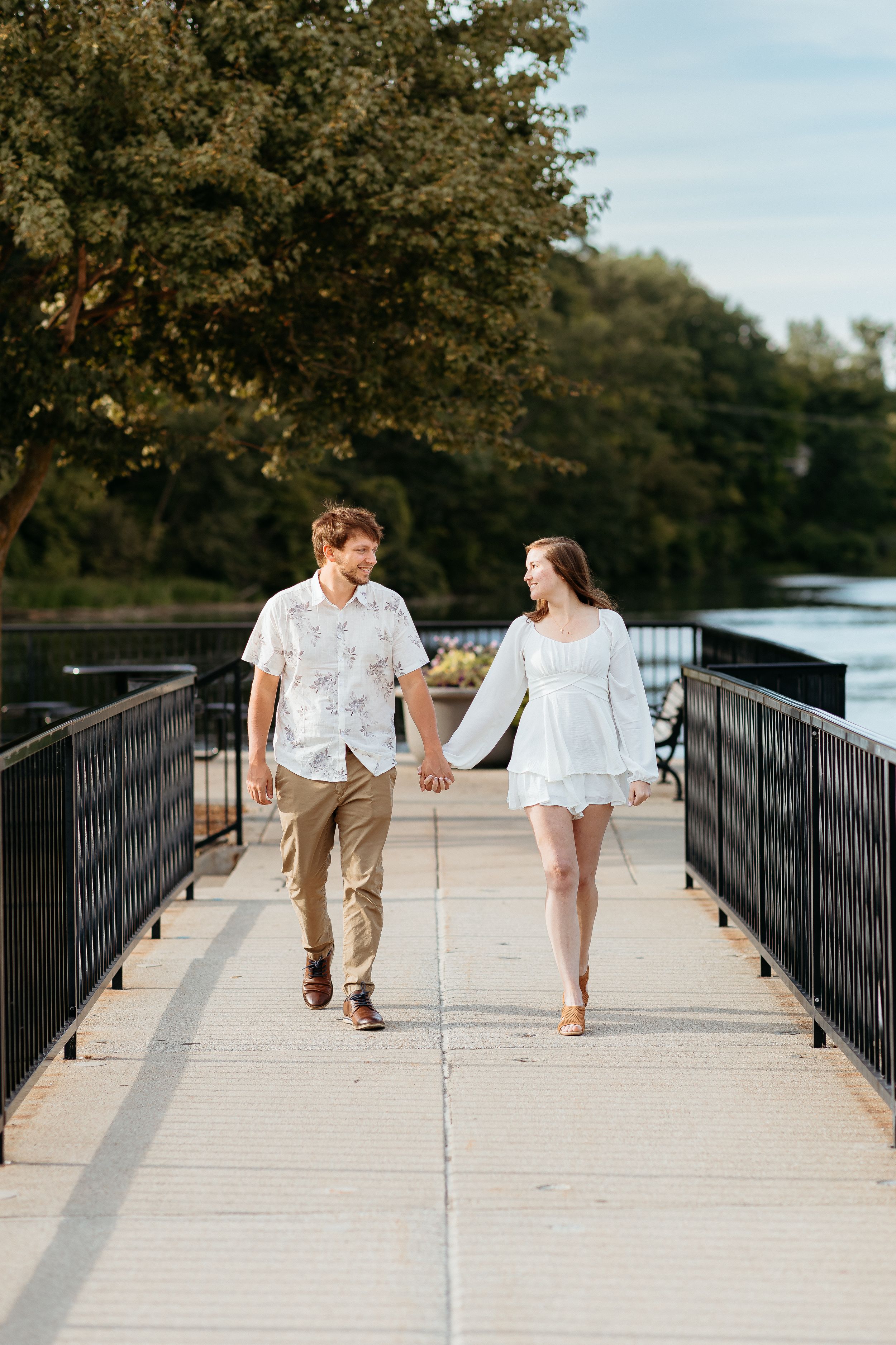 Couple holding hands while walking on waterfront pier in casual summer outfits. Engagement session in Rockford Michigan Summer.