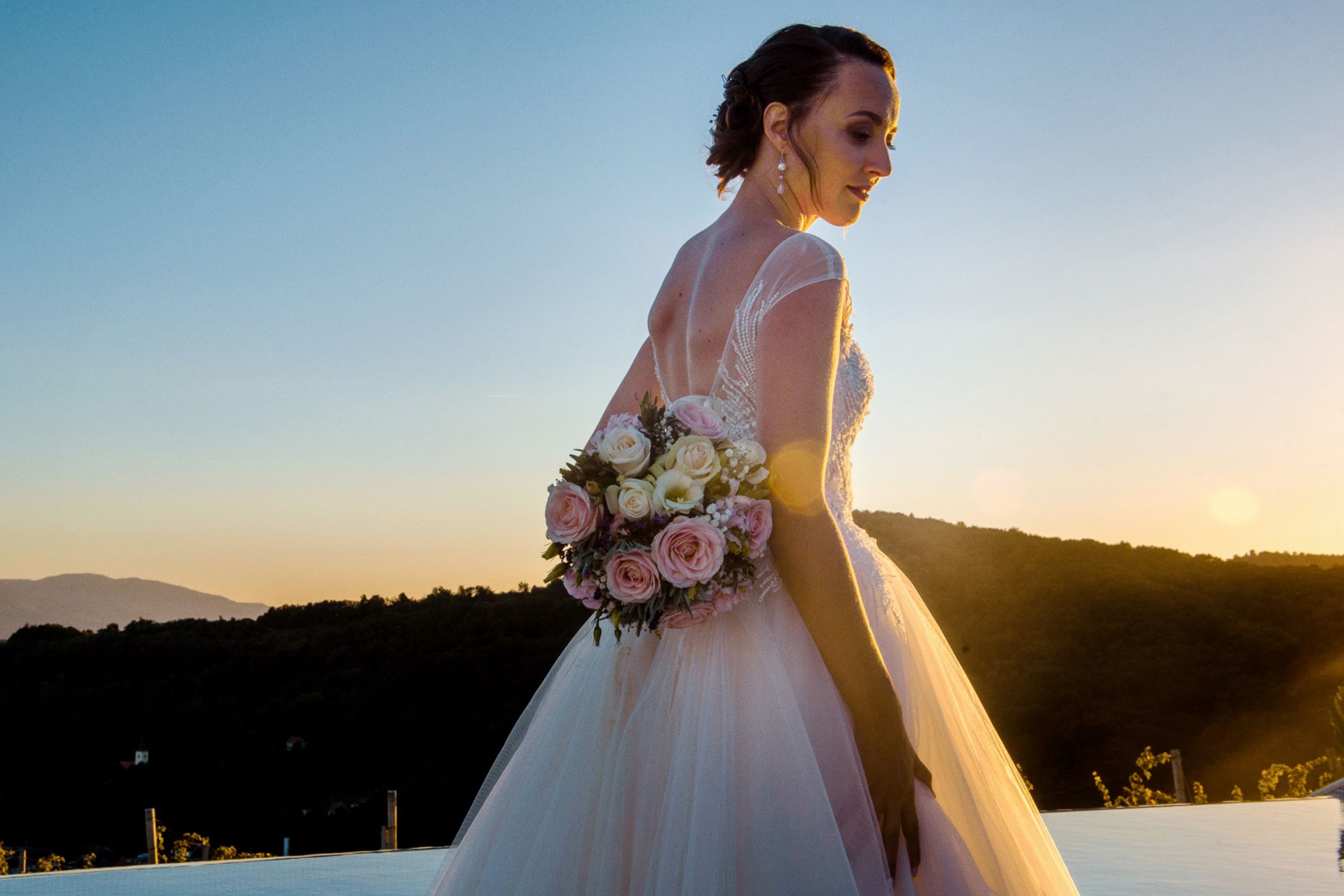 Bride in flowing white gown holds bouquet against mountain sunset. Restavracija Tri Lučke, Krško
