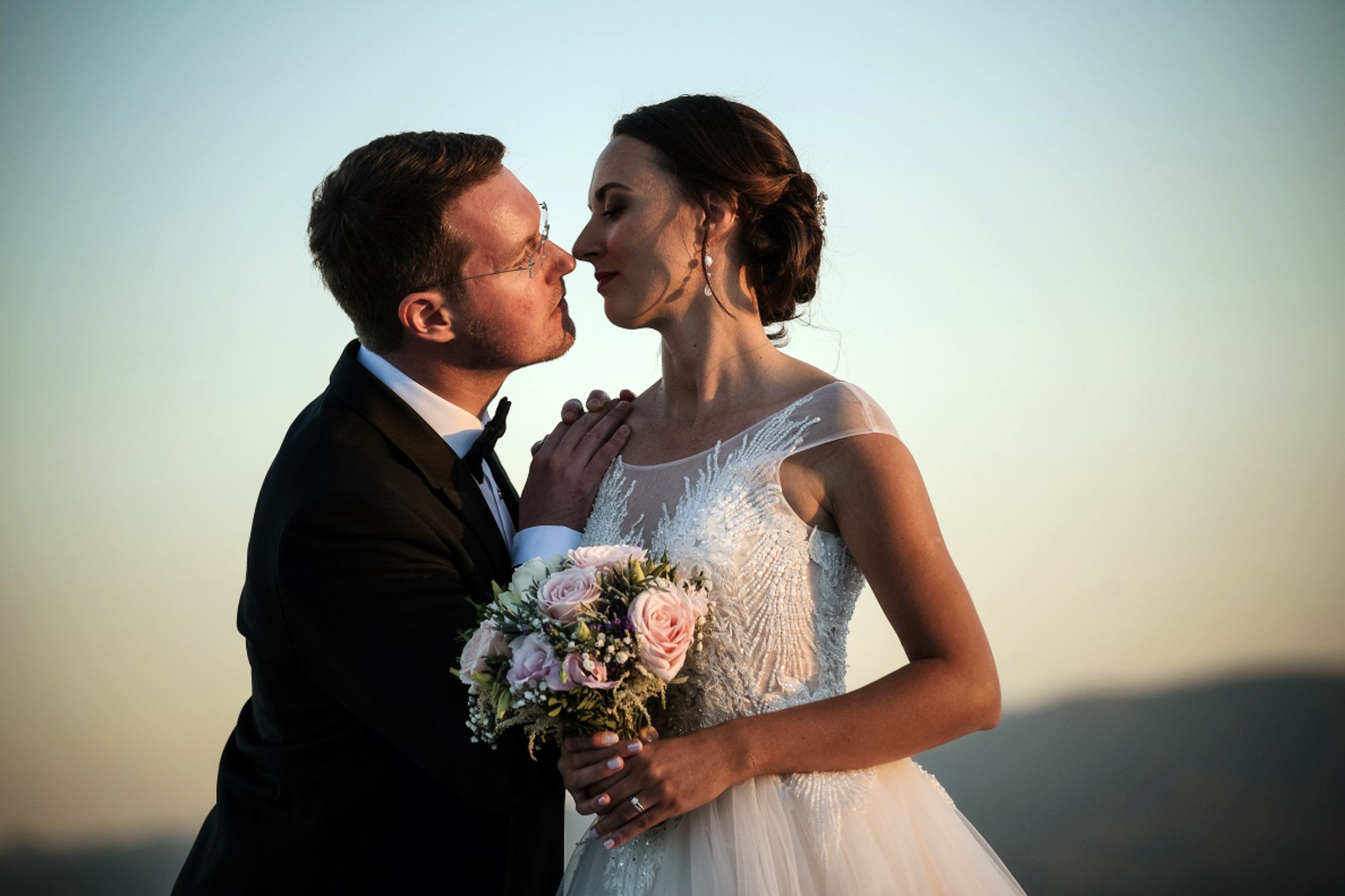 A couple in wedding attire shares romantic kisses during sunset while holding a bouquet.