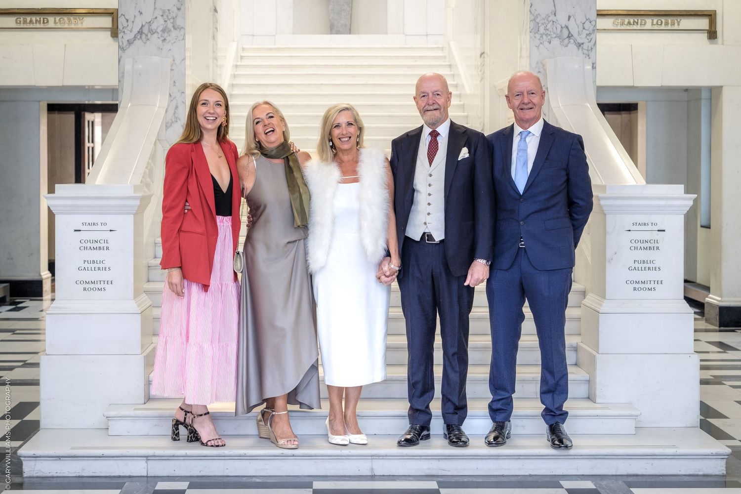 Wedding party members stand together on white marble steps in coordinated formal attire for a group photo.