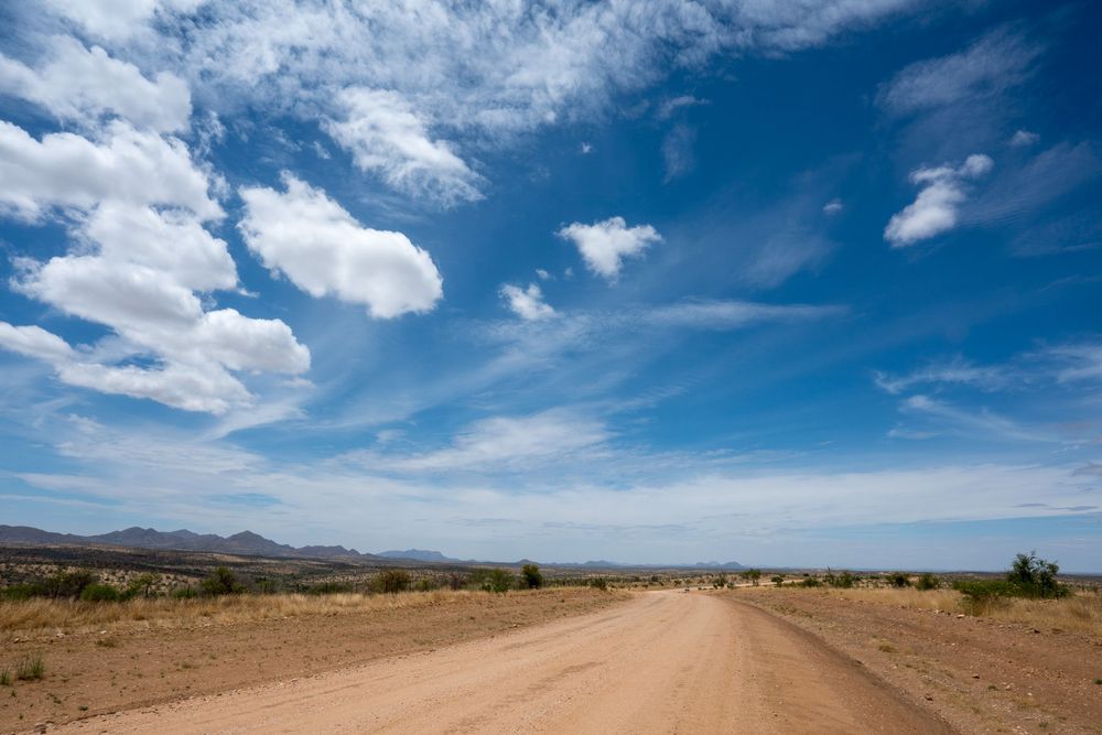 A panoramic landscape shows a winding dirt road through arid terrain under a bright blue sky with scattered white clouds.