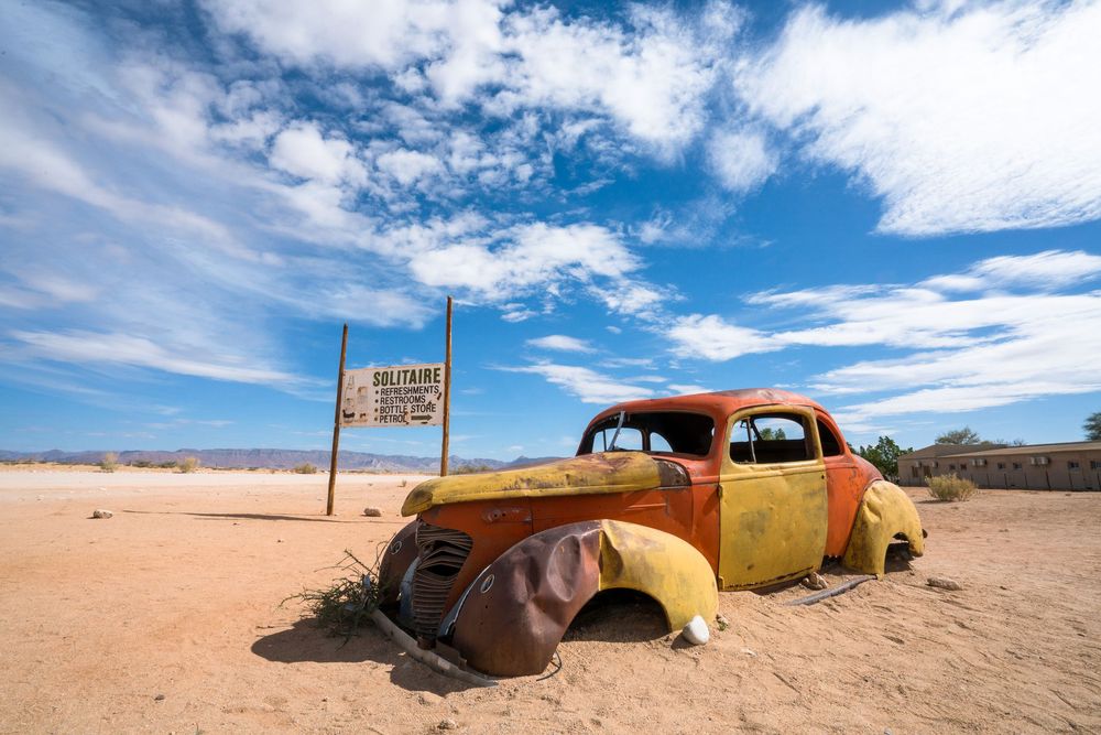 An old abandoned car rusts in the desert sand under blue skies with wispy white clouds stretching to the horizon.