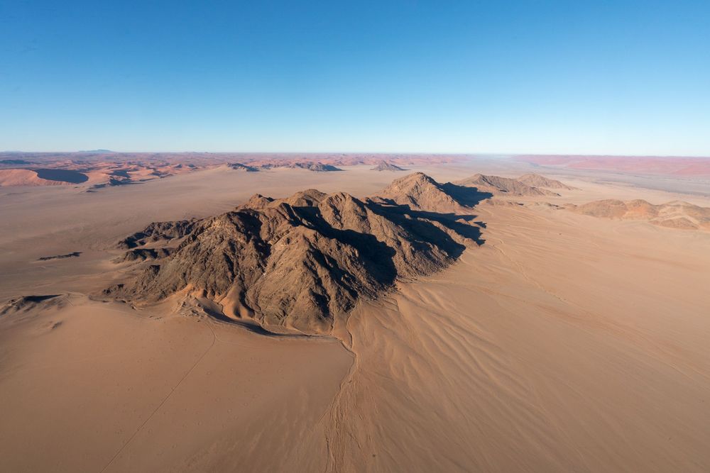 Aerial view of rocky mountains surrounded by vast sand dunes in a desert landscape under clear blue skies.