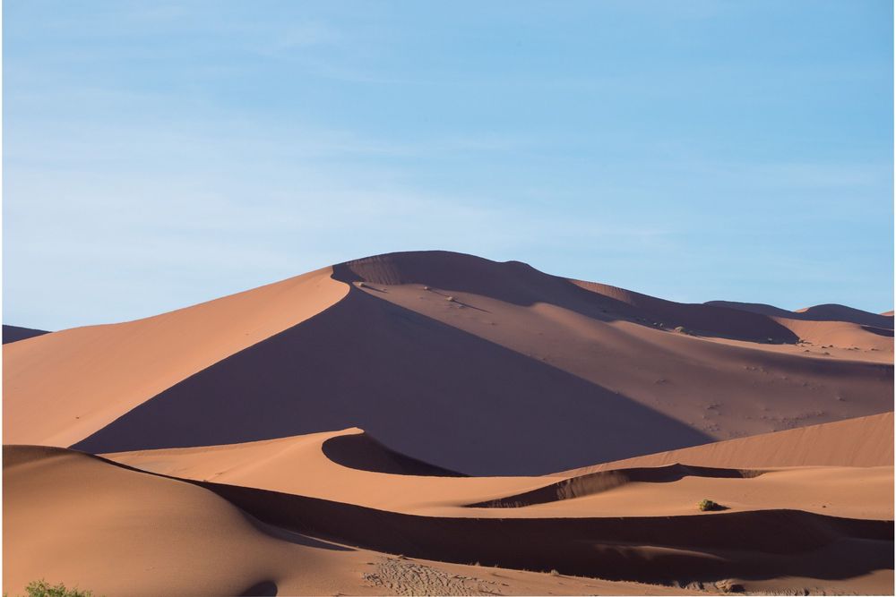 The orange sand dunes of the Namib Desert create a striking pattern against a bright blue sky at sunset.