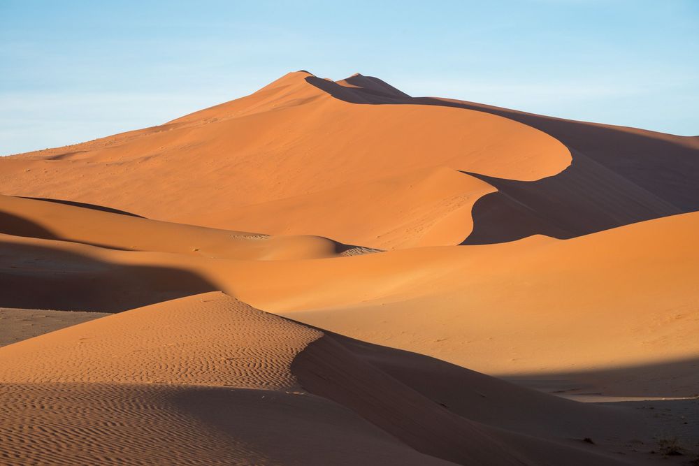 Curved orange sand dunes stretch across a vast desert landscape under bright blue skies.