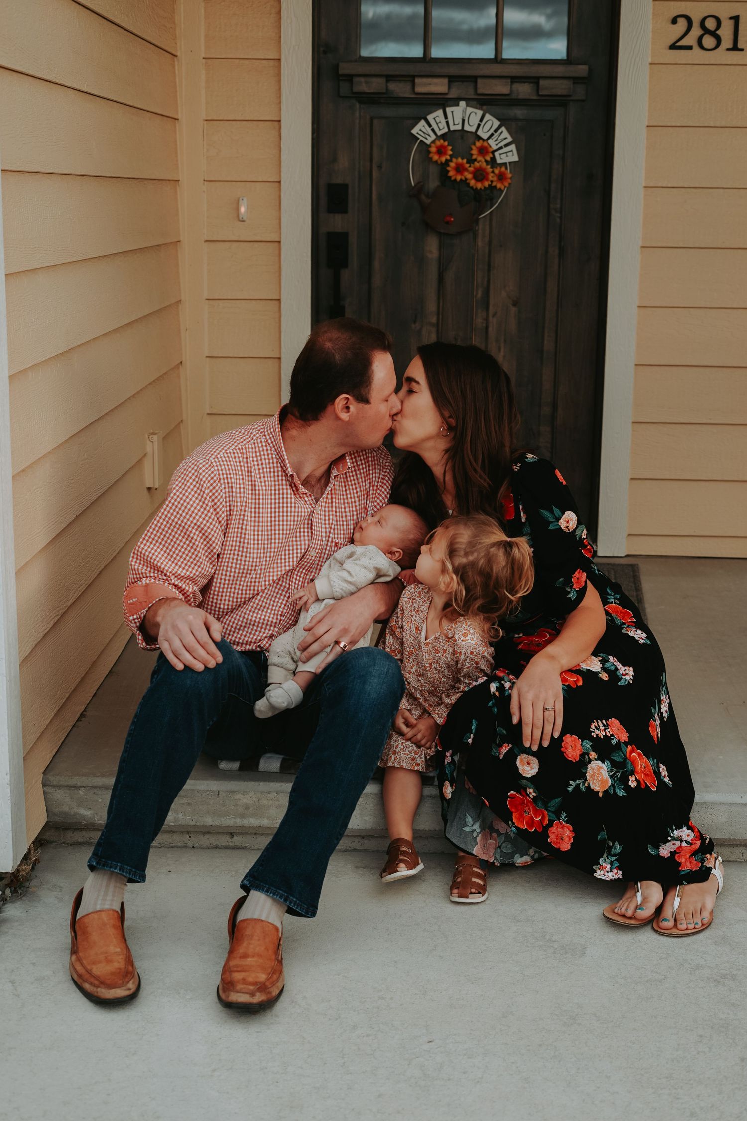 A family sits together on their front porch steps sharing a tender moment outside their home with a seasonal wreath on the door.