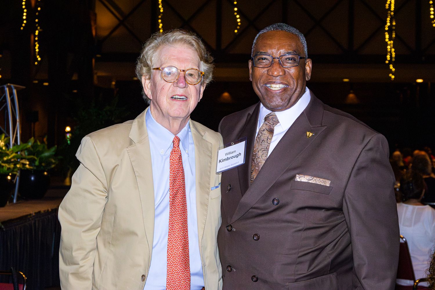 Two people in business attire pose together at an event, one wearing a beige suit with orange tie and one in a brown suit.