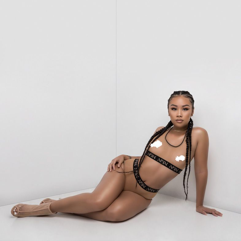 Model wearing black and gold Nude by Nikki lingerie set poses on white background.