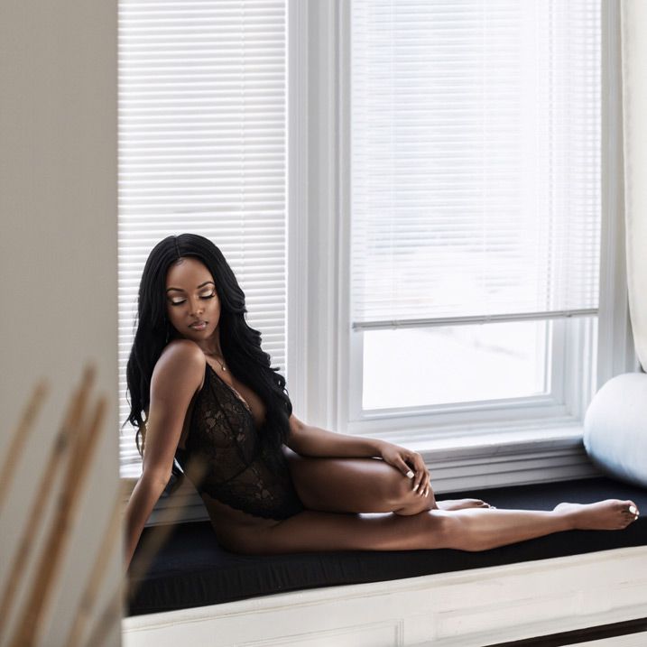 Person in black outfit reclining by window with horizontal blinds in minimalist white room interior.