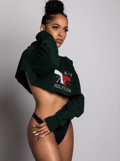 Model wearing cropped Tommy Hilfiger sweatshirt and black bikini bottoms poses against grey background.