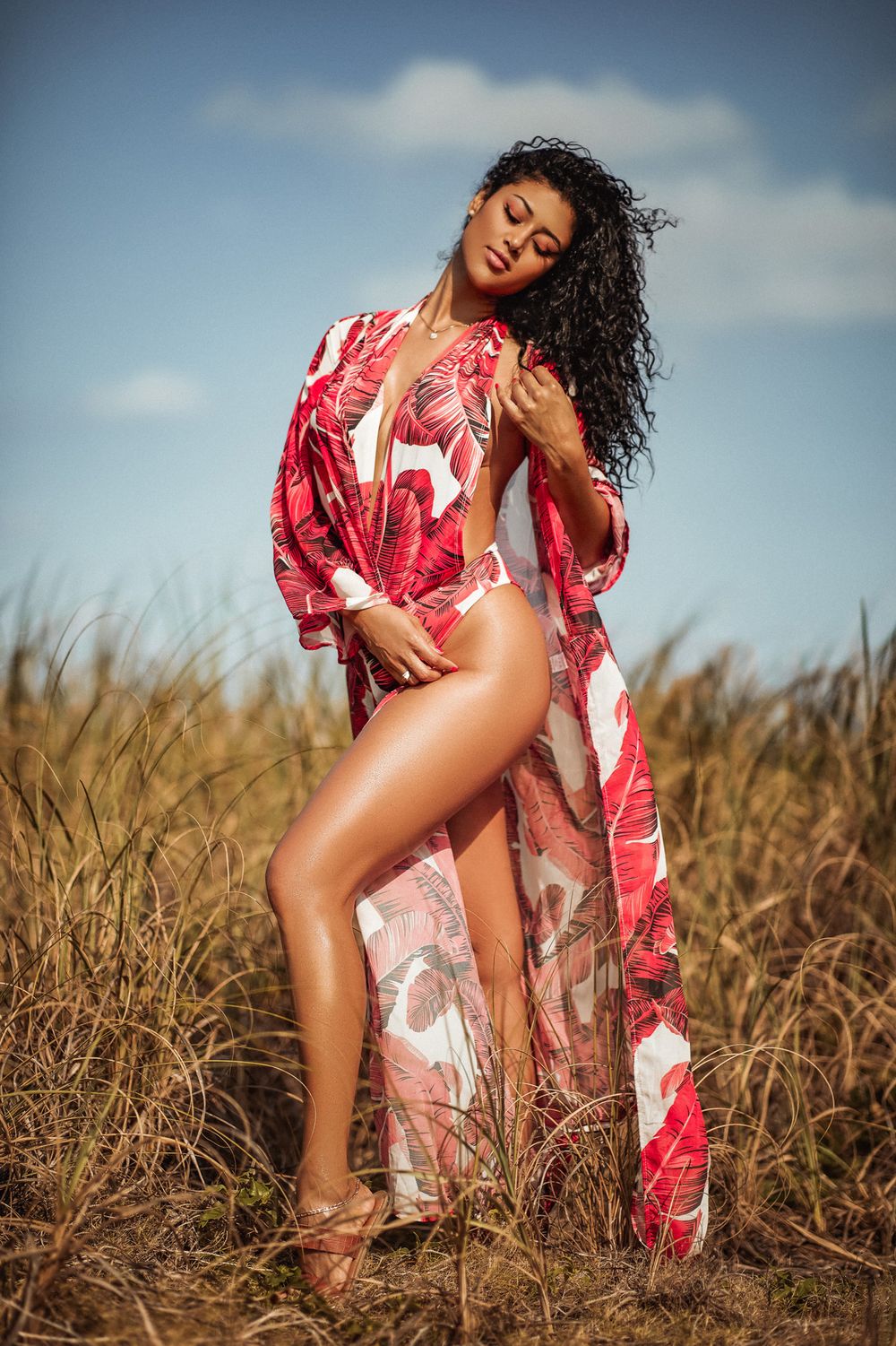 Fashionable beach photoshoot featuring red and white tropical print maxi dress and swimsuit against tall grass and blue sky.