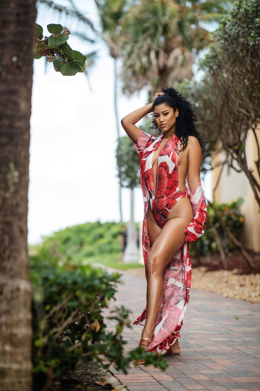 Woman in red and white tropical print swimsuit and matching coverup poses on brick path with palm trees in background.