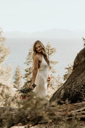 A person in a flowing white dress stands on a scenic cliff overlooking the ocean at sunset among pine trees.