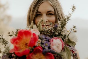 Close-up floral photo showing vibrant coral peonies and purple wildflowers against a soft blurred background in natural light.