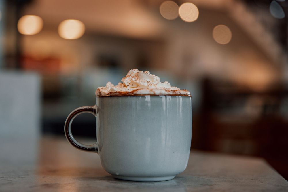 Cup of hot chocolate topped with whipped cream and caramel drizzle in a warm cafe setting with bokeh lights.