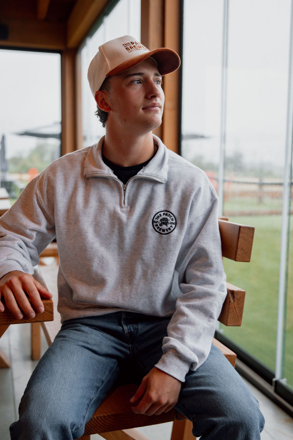 Person in gray sweatshirt and jeans sits by window in minimalist indoor setting.