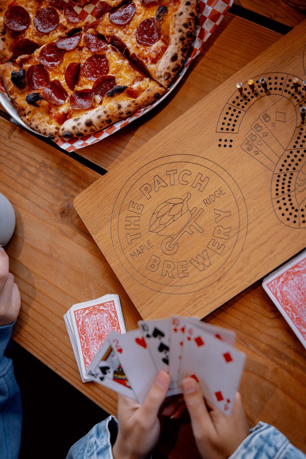 People play cards on a wooden table while sharing pizza at a restaurant called Patch & Brew.
