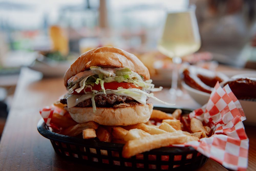 A juicy burger served in a plastic basket with crispy french fries and a glass of white wine in the background.